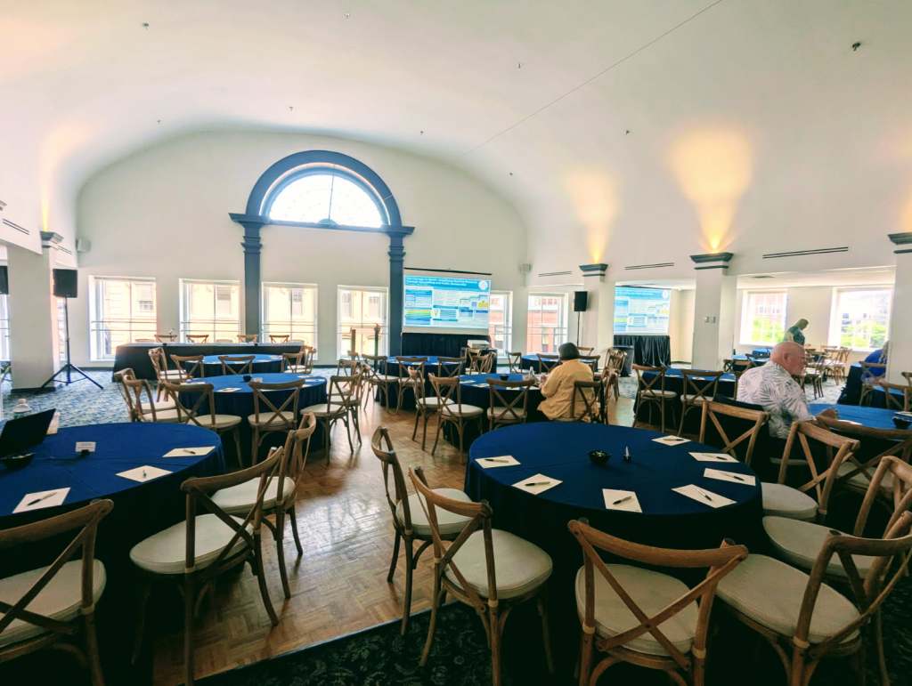 Interior view of a conference room set up for the INANE 2025 event, featuring round tables with blue tablecloths, chairs, and a large screen displaying information.