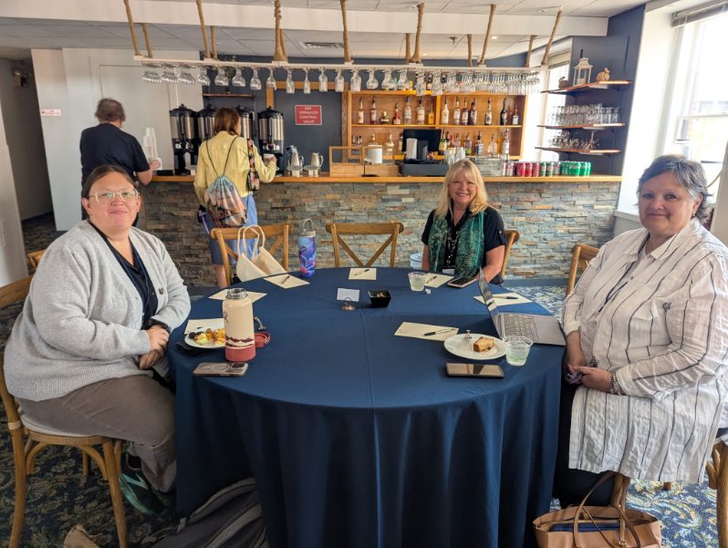 A group of three women seated at a round table covered with a blue tablecloth, with snacks and a laptop present. A bar area is visible in the background.
