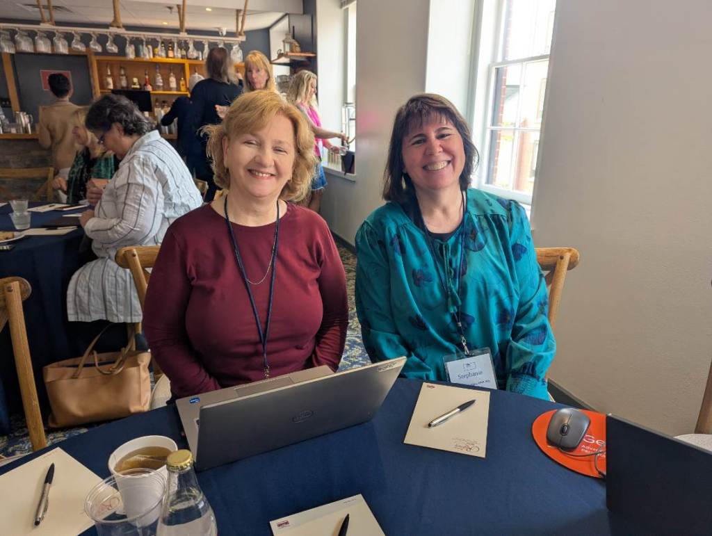 Two women smiling and sitting at a table during the INANE 2025 conference, with laptops and notepads in front of them.