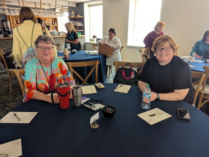 Two women sitting at a table during the INANE Conference, with one woman wearing a colorful top and holding a drink, and the other wearing a black shirt. The room has other attendees in the background.