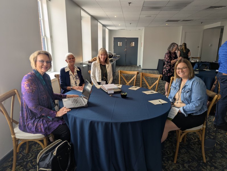 A group of four women seated at a round table during the INANE Conference, engaging in discussion with a laptop and notepads present.