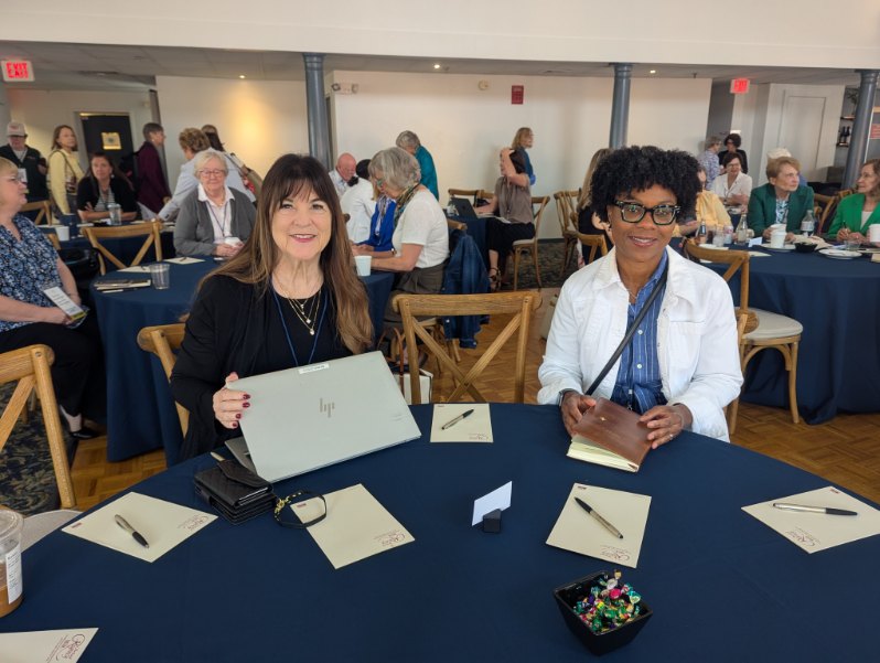Two women sitting at a round table during the INANE 2025 conference, each with a laptop and notebooks, with a gathering of attendees in the background.