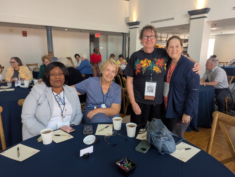 Four women sitting at a table during a conference, with coffee cups and materials in front of them, engaging in conversation.