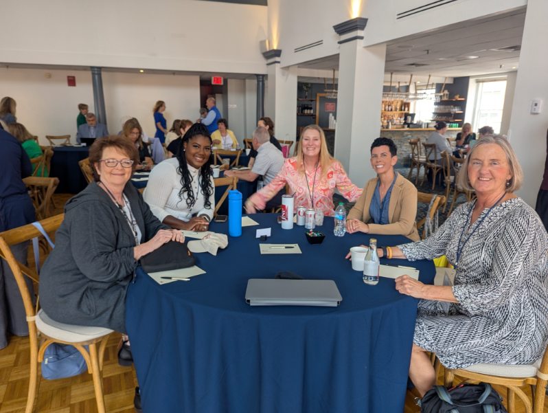 A group of five women seated at a round table with a blue tablecloth during the INANE 2025 conference, engaging and smiling. The background shows other conference attendees and a bar area.