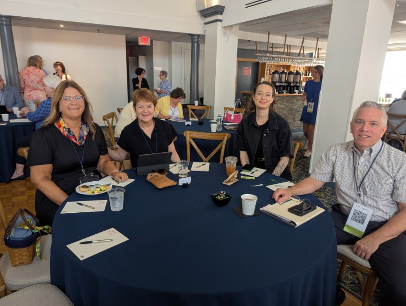 Four attendees sitting at a round table during the INANE Conference, with food and drinks in front of them.