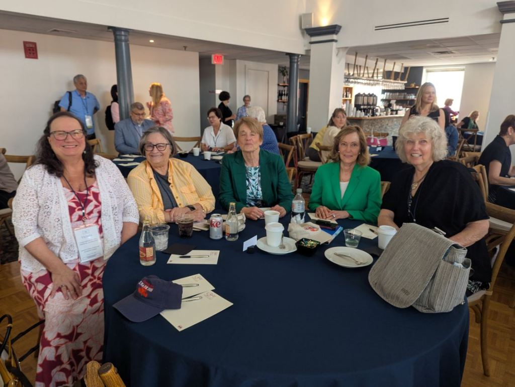 Five women seated at a round table with a dark tablecloth, engaged in conversation during the INANE 2025 conference. They are smiling and appear to be enjoying the event, with various drinks and plates on the table.