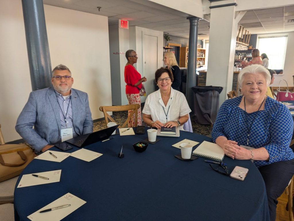 Three conference attendees seated at a round table with beverages and notebooks, engaged in conversation. The setting appears to be a casual meeting space.