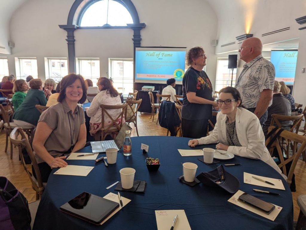 A group of conference attendees seated at tables in a well-lit venue, engaging in conversation with a presentation screen displaying 'Hall of Fame 2023' in the background.