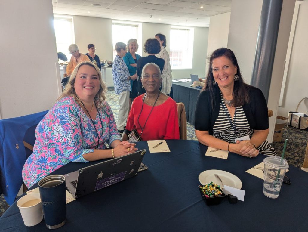 Three women smiling at a table during the INANE 2025 conference, with attendees in the background.