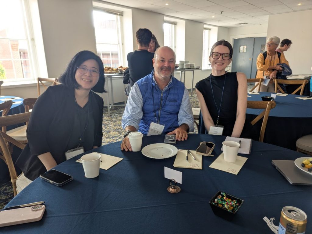 Three attendees at a conference seated at a table, smiling and engaging. The table is set with coffee cups and notebooks, with a background of a well-lit room.