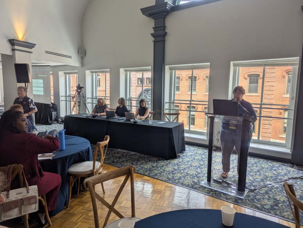 A conference setting with a speaker at a podium, presenting to an audience seated at round tables. A panel of speakers is visible at the back, with large windows showing a brick building outside.
