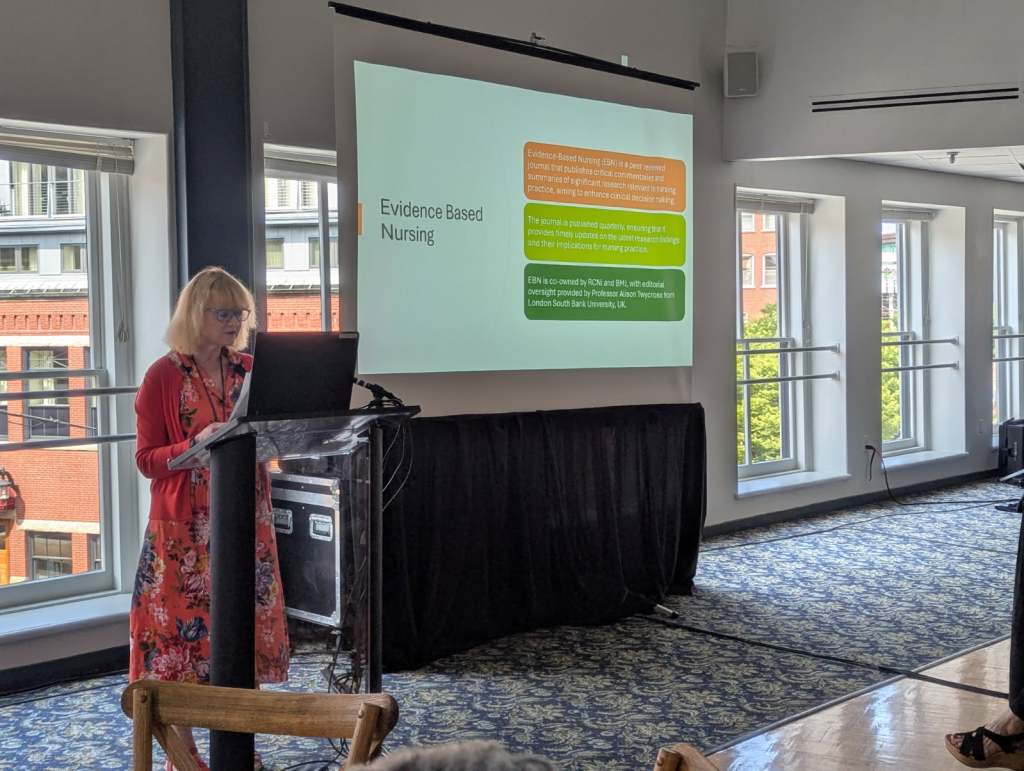 A woman presenting at a conference, standing in front of a projector screen displaying information about the Evidence Based Nursing journal.