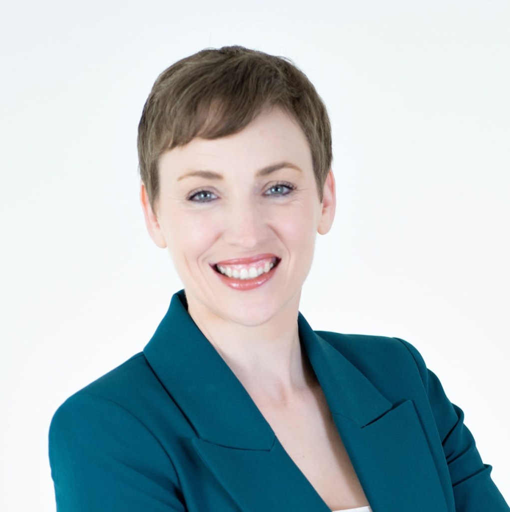 Portrait of a smiling woman with short hair, wearing a teal blazer, against a plain white background.