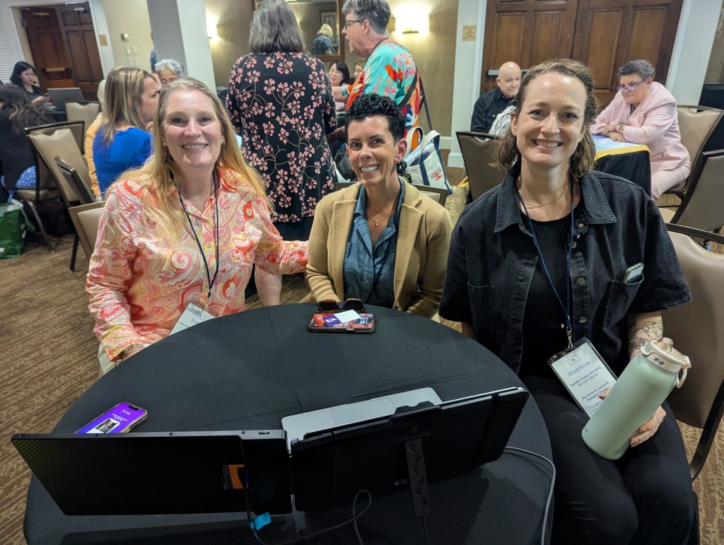 Three women posing at a conference table with a laptop and water bottle, smiling at the camera.