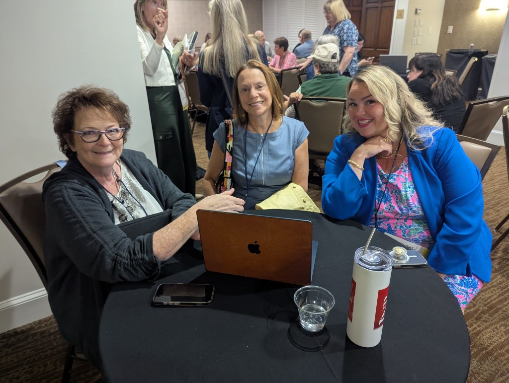 Three women seated at a round black table during a conference, smiling and engaging with a laptop and drinks. The background features other attendees in a meeting space.
