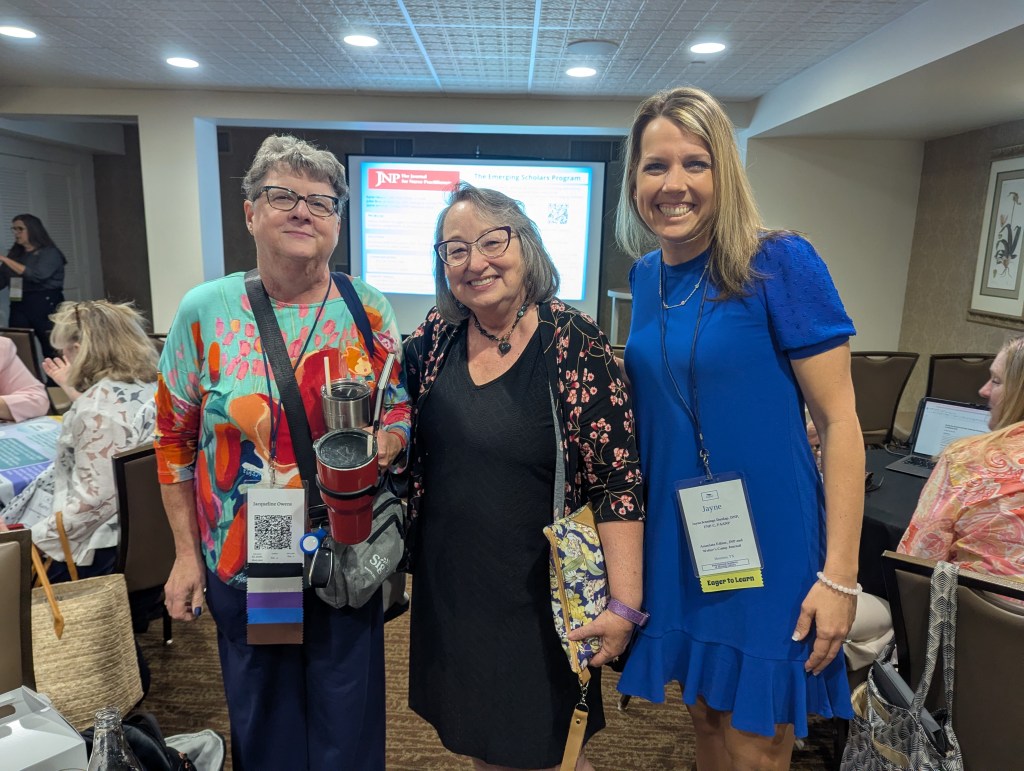 Three women posing together at a conference, with a background of people and presentation materials. One woman wears a colorful shirt, another wears a black outfit, and the last wears a blue dress.
