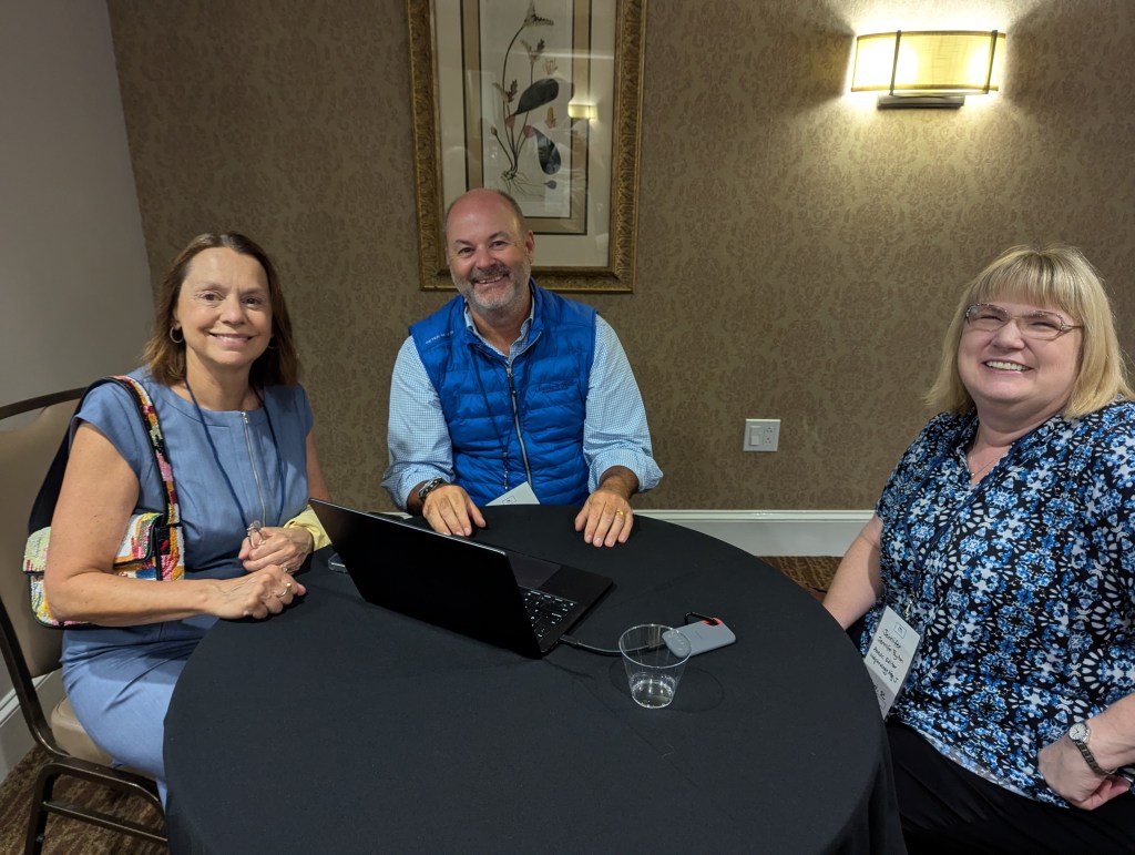 Three people seated at a black table during a conference, engaged in conversation. One person has a laptop open in front of them, with a glass of water beside it. The background features patterned wallpaper and a piece of framed artwork.