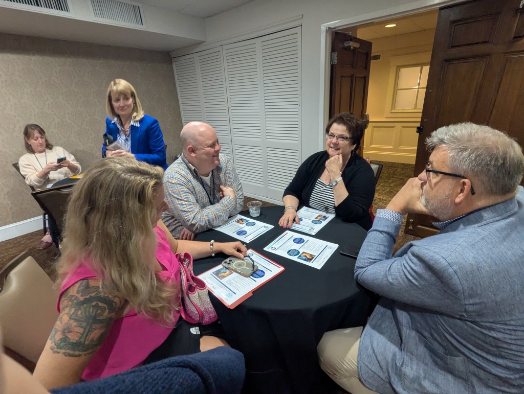 A group of five people engaged in conversation at a table during a conference, with materials and drinks on the table. One person is seated, looking at papers while others are interacting.