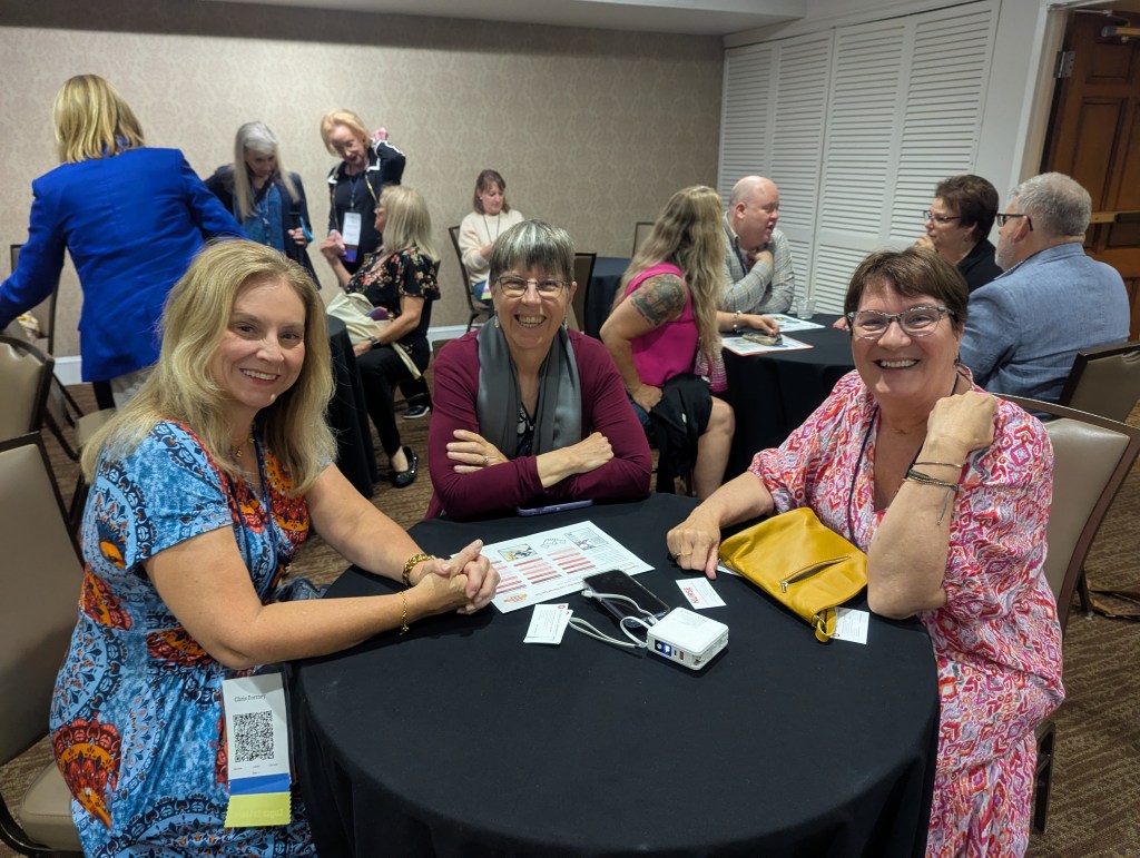 Three women smiling around a table during a conference, with others in the background engaging in conversation.