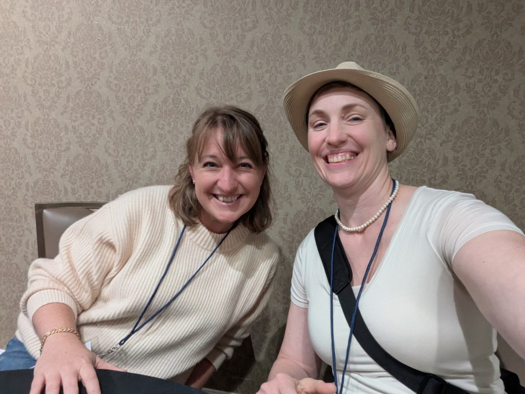 Two women smiling for a selfie in a conference setting, with a patterned wall in the background.