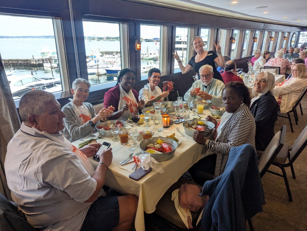 A group of diners enjoying a festive lobster dinner at a restaurant with views of a harbor, with some people cracking lobster shells and others smiling at the table.