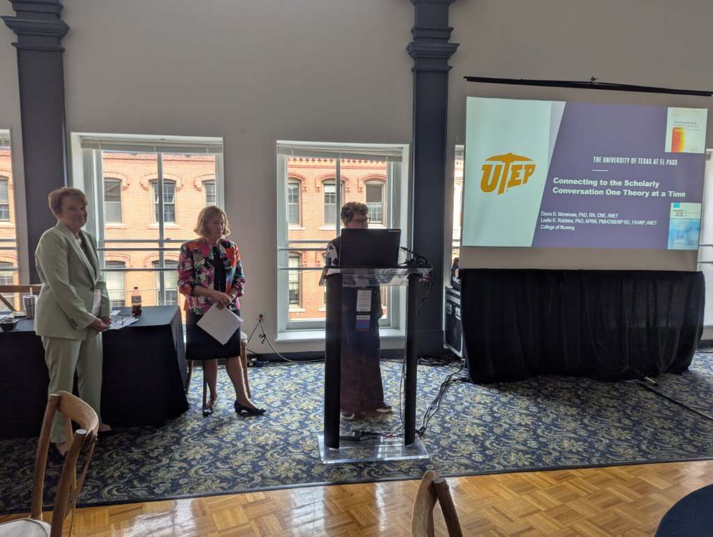 Three speakers at a conference presentation, with one presenting at a podium and two others standing to the side. The backdrop includes a screen displaying the title of the presentation related to scholarly conversations and nursing theory.