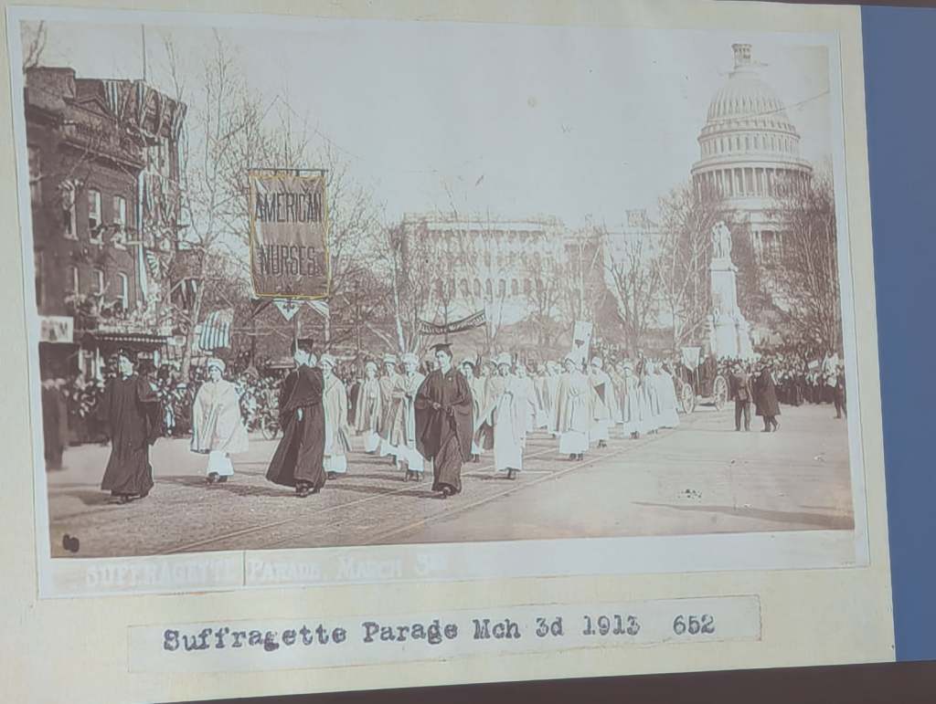 Historic photograph of the Suffragette Parade on March 3, 1913, showcasing a procession of women dressed in white, carrying banners, with the U.S. Capitol building in the background.