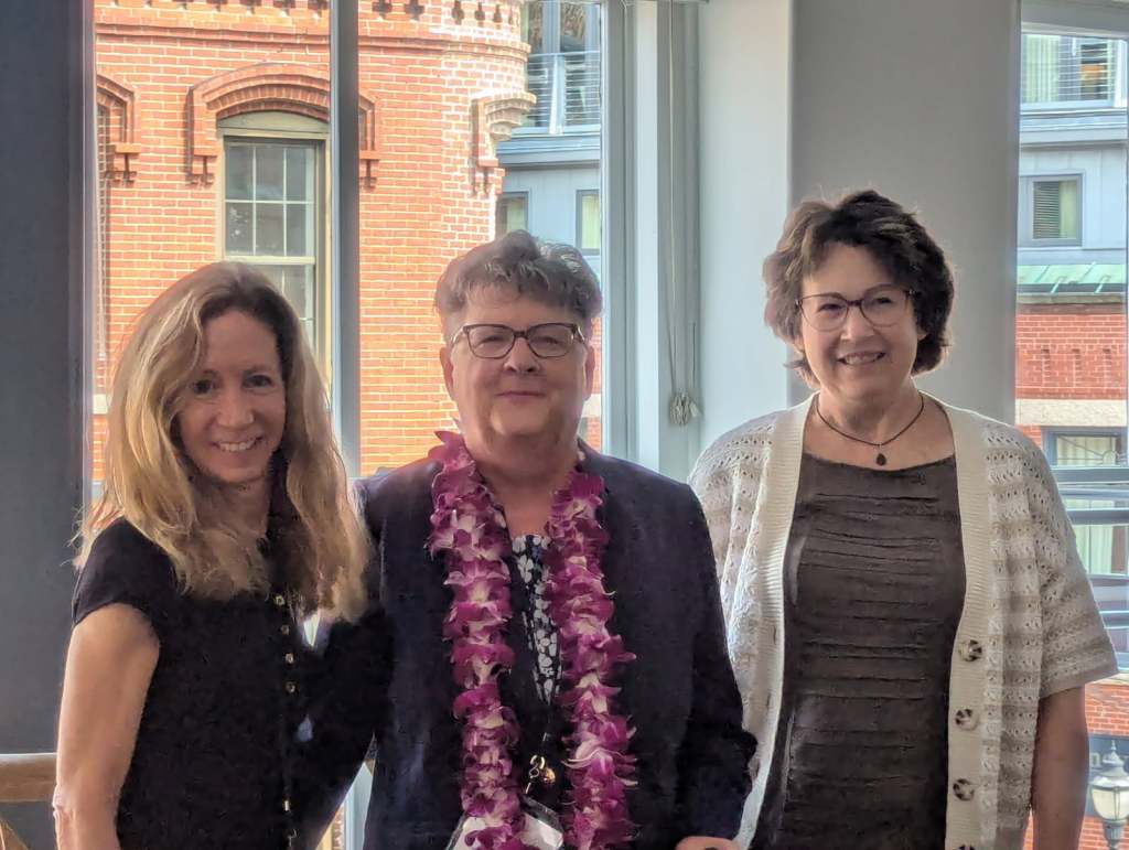 Three women posing together at a conference, with a brick building visible in the background. One woman is wearing a floral lei.