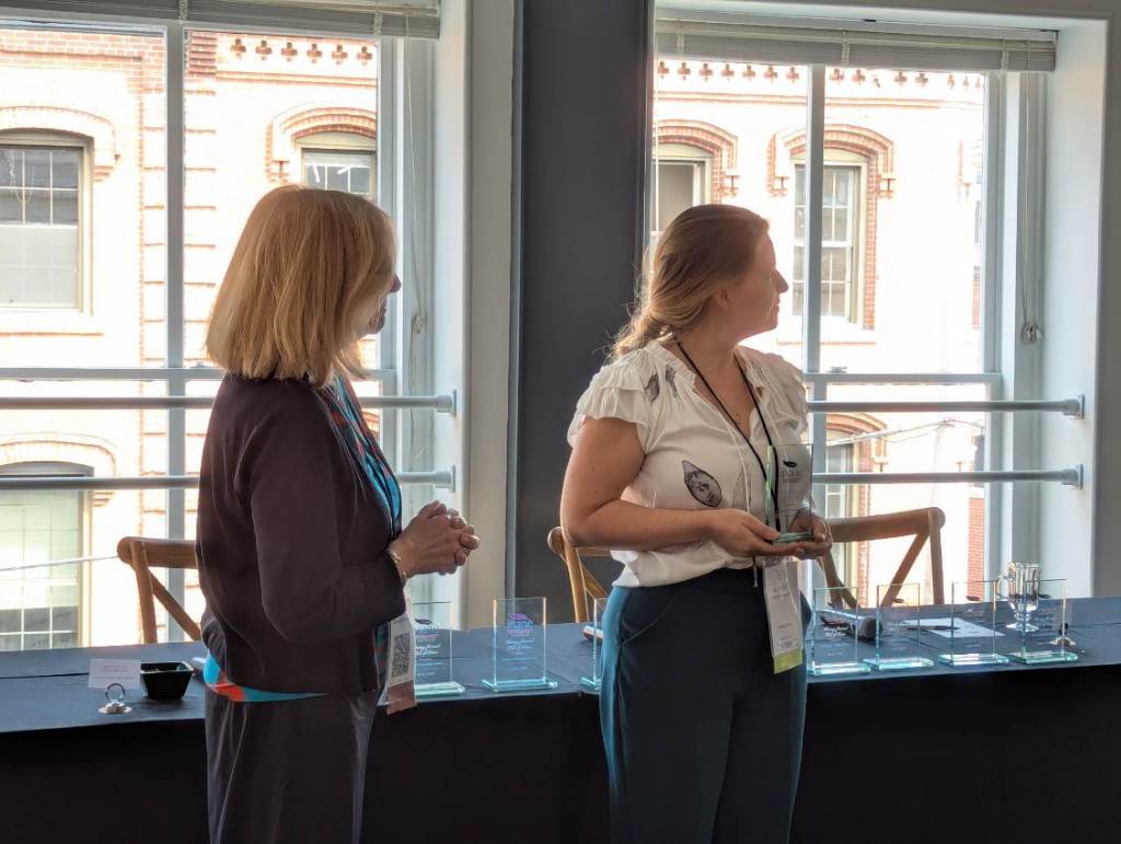 Two women stand near a table displaying awards and trophies, engaged in conversation during the Nursing Journal Hall of Fame Ceremony.