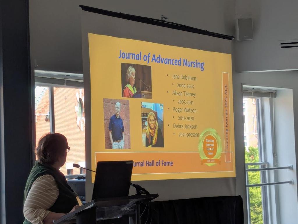 A presenter stands at a podium facing a large screen displaying the Nursing Journal Hall of Fame with names and years of recognition for various individuals.