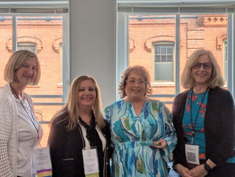 Four women standing together in an indoor setting, smiling at the camera. The background features large windows with a brick building visible outside. Each woman is wearing a name tag.
