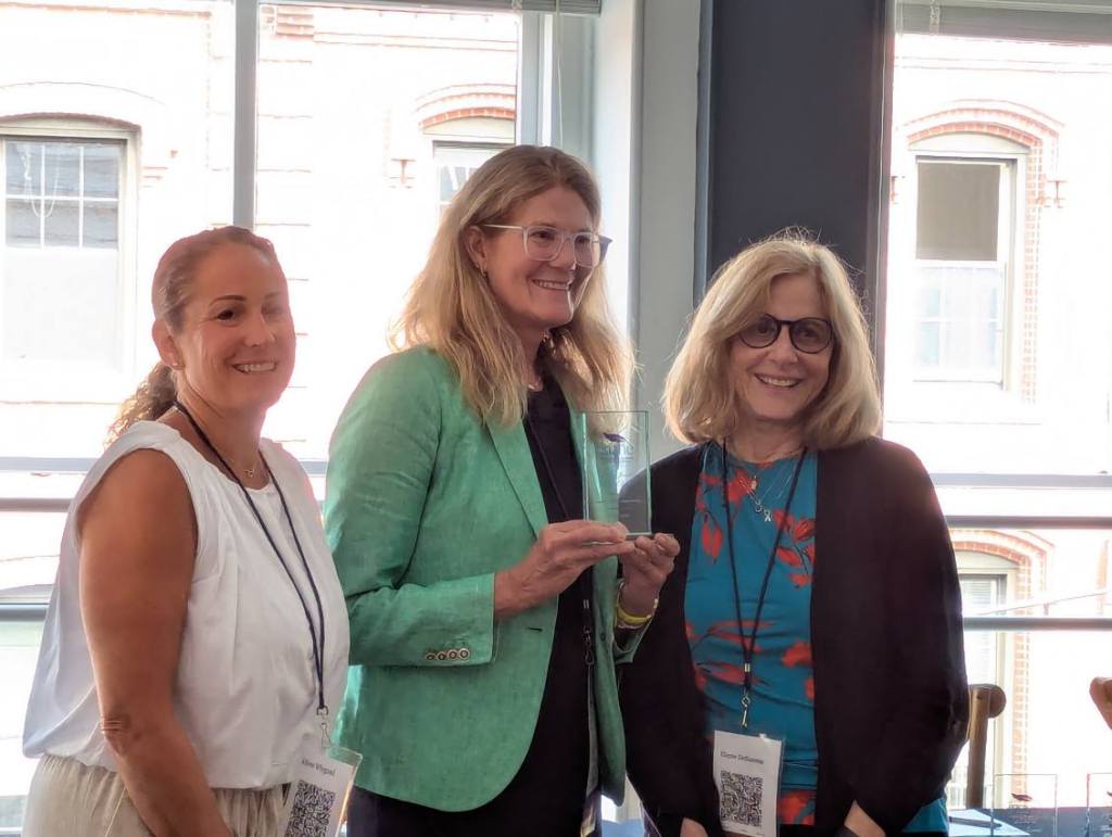 Three women stand together in a well-lit room, smiling while one of them holds an award. The woman on the left has a ponytail and wears a white top, while the second woman in a green blazer is presenting the award, and the third woman on the right is wearing glasses and a black cardigan.