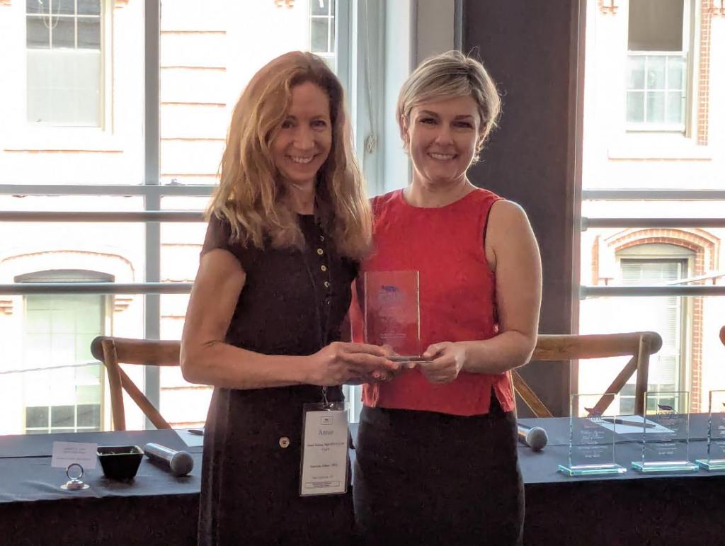 Two women smiling and holding an award in front of a table with several awards displayed. The background features large windows with a view of the outside.