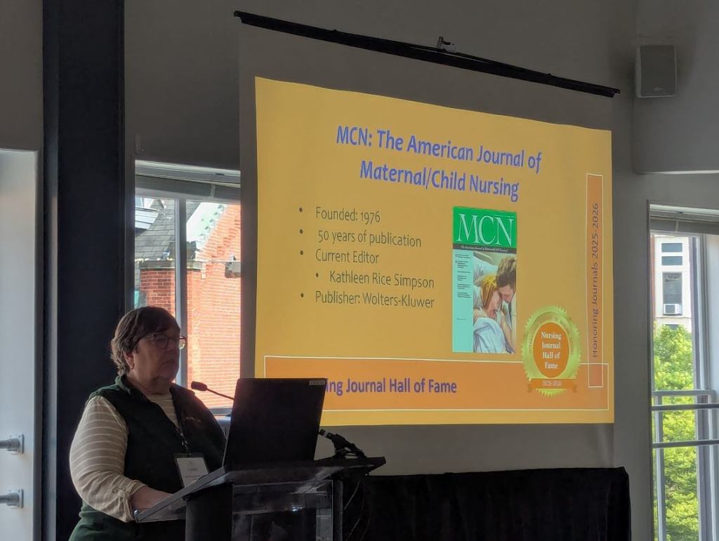 A presenter stands at a podium speaking at the Nursing Journal Hall of Fame Ceremony, with a large screen behind displaying information about the MCN: The American Journal of Maternal/Child Nursing.