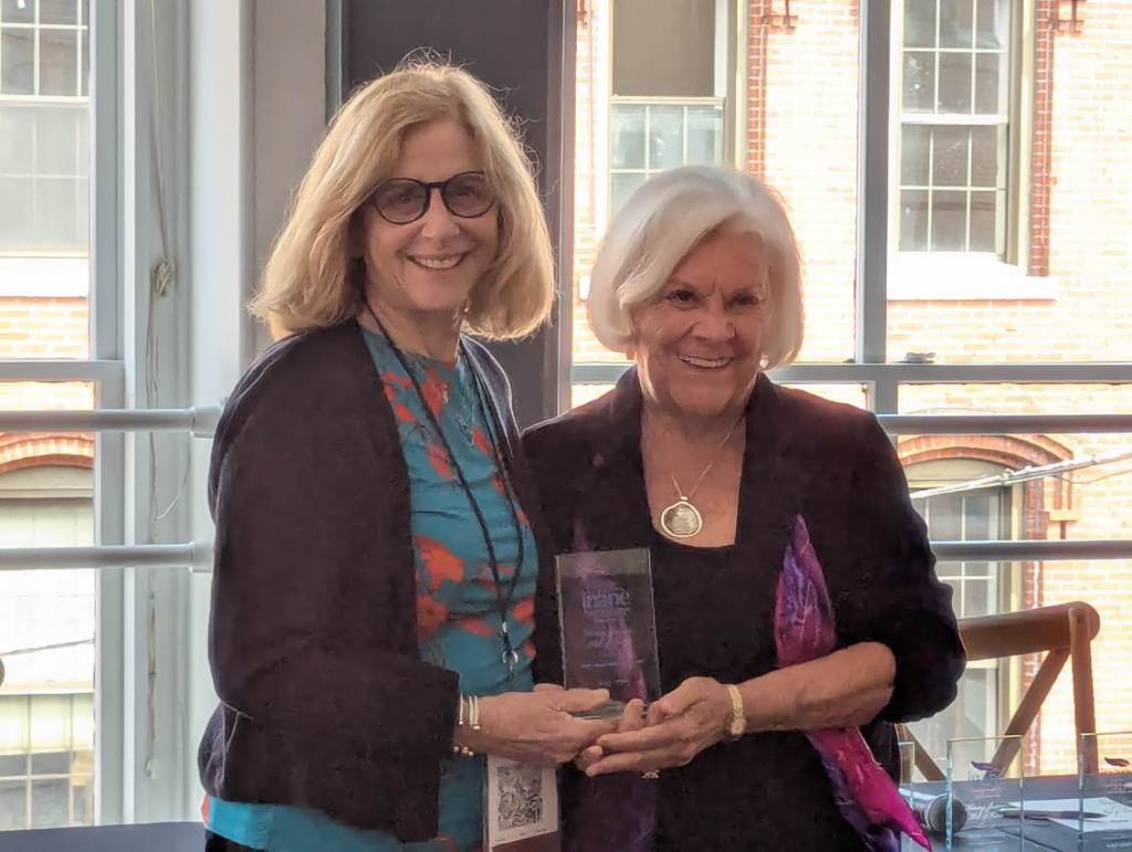 Two women smiling while holding an award at the Nursing Journal Hall of Fame Ceremony, with large windows in the background.