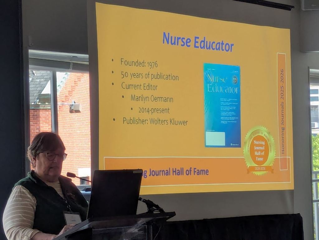 A woman presenting at a conference, speaking about the Nurse Educator journal. A slide in the background lists details such as its founding year, current editor, and publisher.