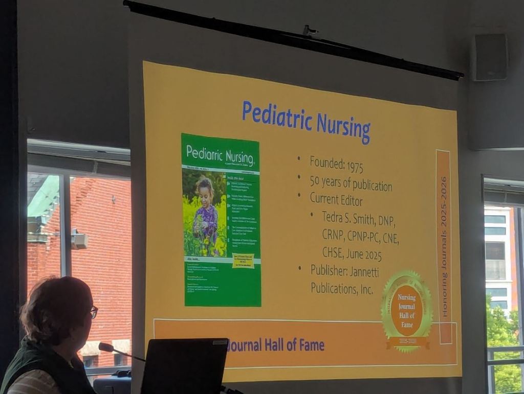 A woman wearing glasses sits in front of a microphone, looking at a presentation about the Pediatric Nursing journal, which highlights its founding date, current editor, and publication details. The presentation includes a colorful cover image featuring a child in a field of flowers.