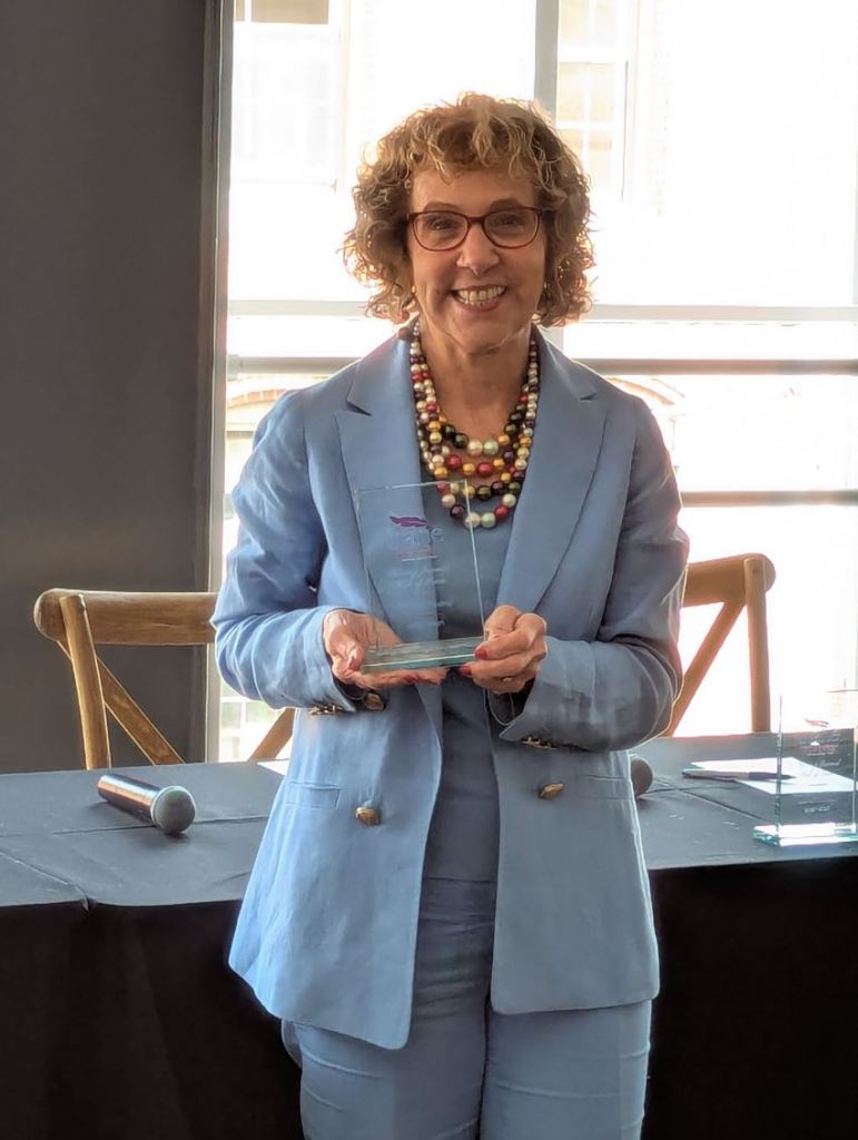 A smiling woman with curly hair and glasses, wearing a light blue suit, holds an award while standing in front of a window.