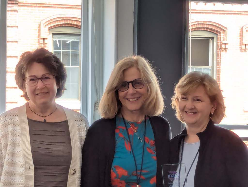 Three women smiling together at the Nursing Journal Hall of Fame Ceremony, with a trophy in one woman's hands and large windows in the background.