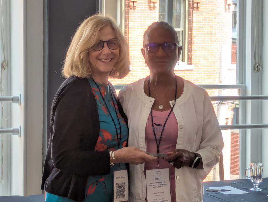 Two women smiling and posing together during the Nursing Journal Hall of Fame Ceremony, one holding an award or recognition, with a bright interior and views of a brick building in the background.
