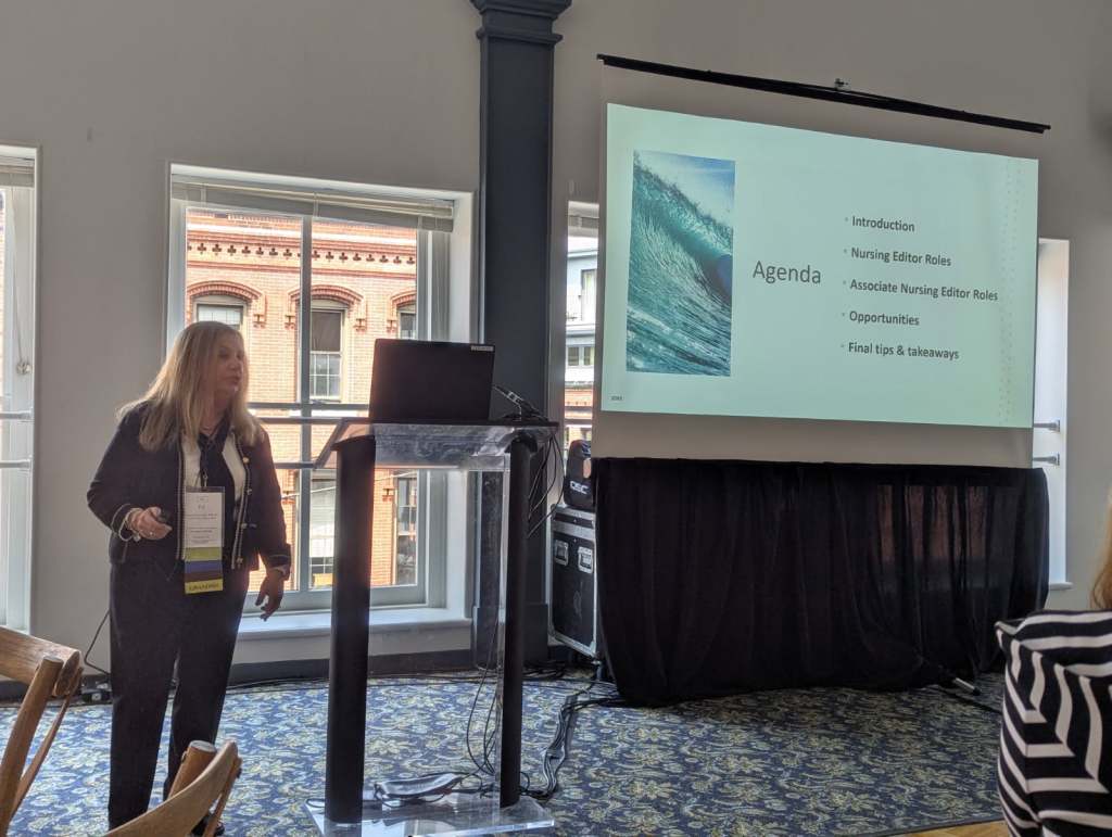 A speaker presenting at the INANE conference with a laptop and agenda displayed on a screen, highlighting topics such as Nursing Editor Roles and opportunities, in a conference room setting.