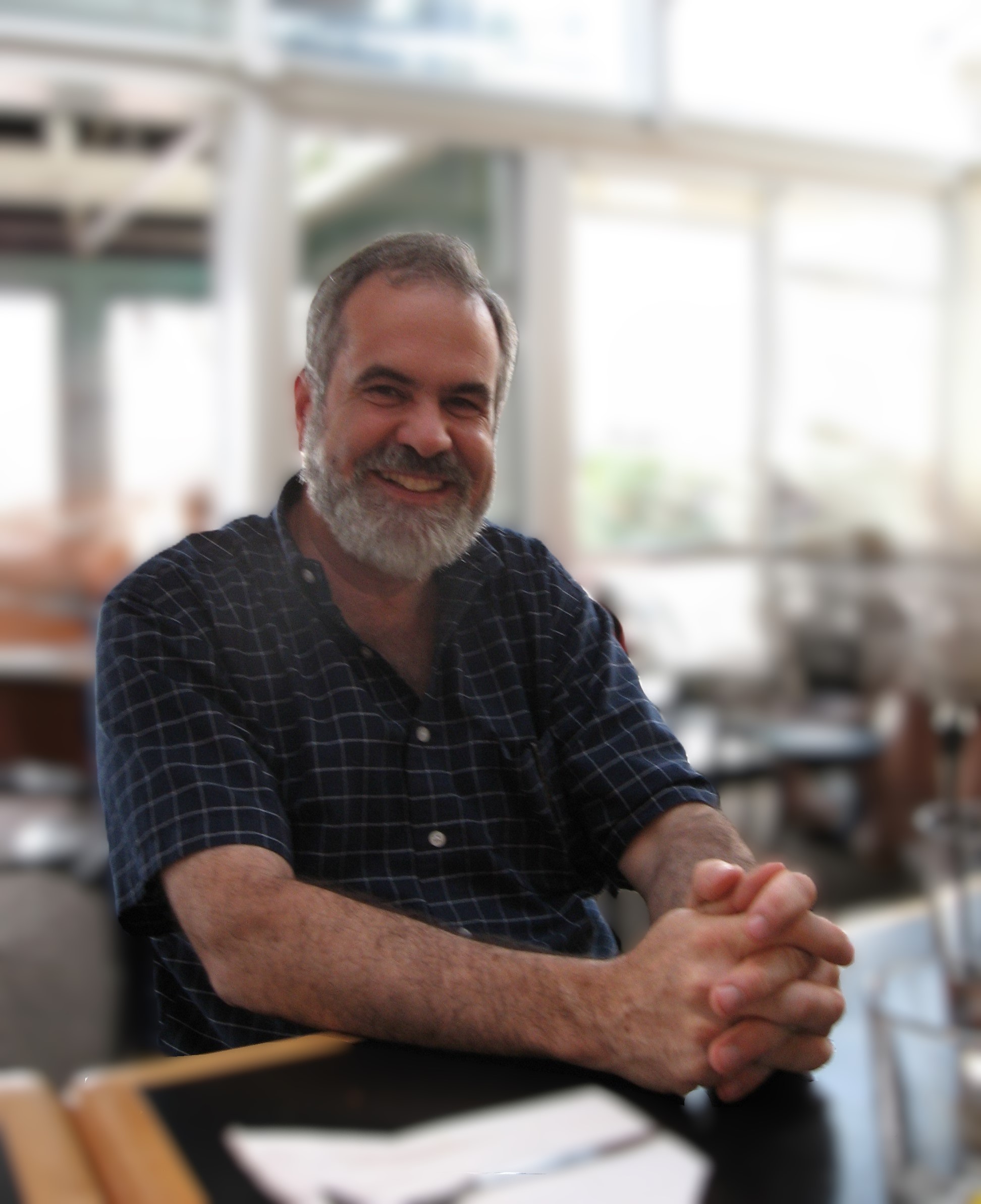 A smiling man with a gray beard sits with his hands clasped in front of him, wearing a blue plaid shirt, in a bright, airy indoor space.