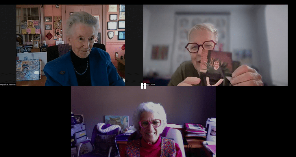 Three older women participating in a video call, with one holding a photo. The background includes art and memorabilia.