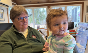 An older woman wearing glasses sits in front of a computer while holding a young child with curly hair, both looking thoughtfully in different directions. The setting appears to be a cozy room with natural light.